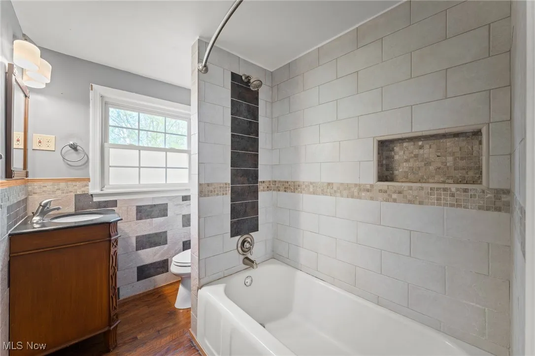 Full bathroom with tile walls, dark wood-style floors, shower / washtub combination, a wainscoted wall, and vanity