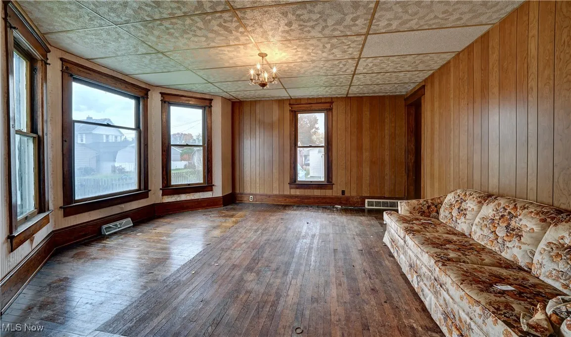 Unfurnished living room featuring dark wood finished floors, wooden walls, a drop ceiling, and a chandelier
