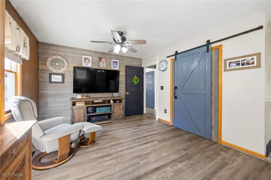 Living area with wooden walls, a barn door, a ceiling fan, an accent wall, and wood finished floors