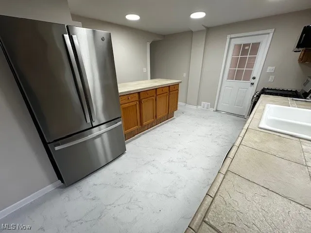 Kitchen with freestanding refrigerator, brown cabinets, tile counters, gas range, and recessed lighting