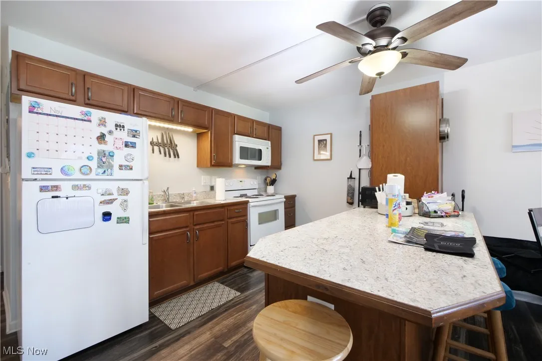 Kitchen featuring light countertops, white appliances, brown cabinetry, dark wood-style flooring, and a kitchen bar