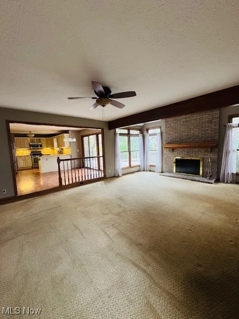 Unfurnished living room featuring a textured ceiling, healthy amount of natural light, a brick fireplace, light carpet, and a ceiling fan