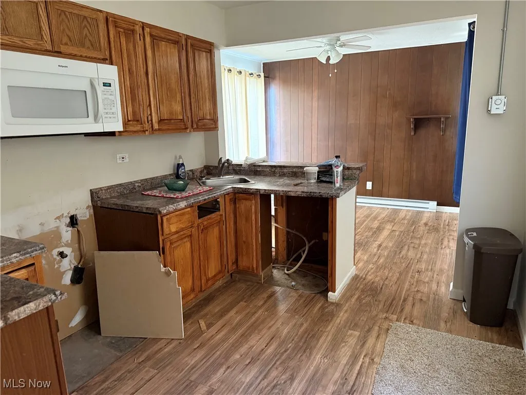 Kitchen with brown cabinetry, white microwave, dark wood-style floors, dark countertops, and wood walls