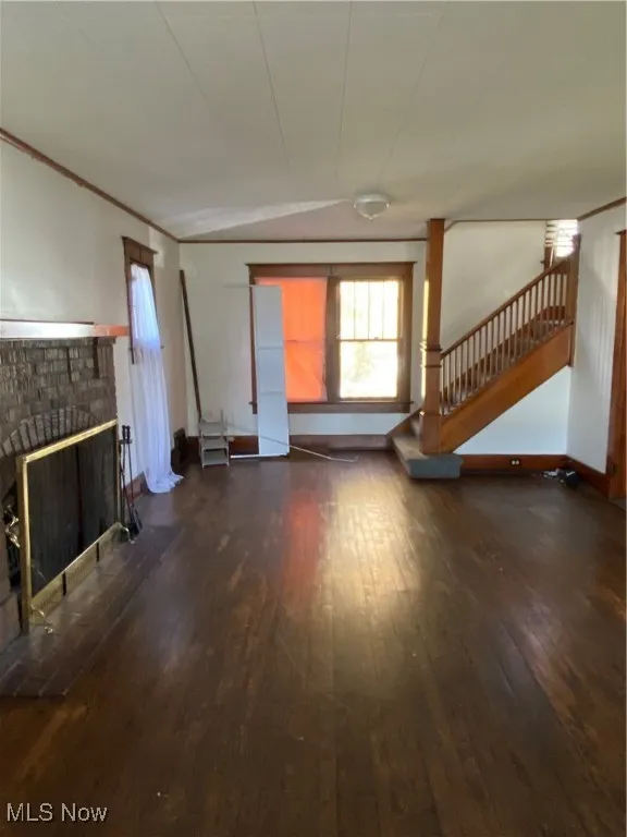 Unfurnished living room featuring dark wood-style floors, stairs, a fireplace, and crown molding