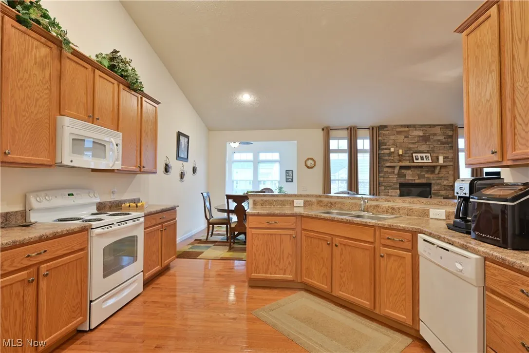 Kitchen featuring white appliances, light wood-type flooring, lofted ceiling, a stone fireplace, and a peninsula