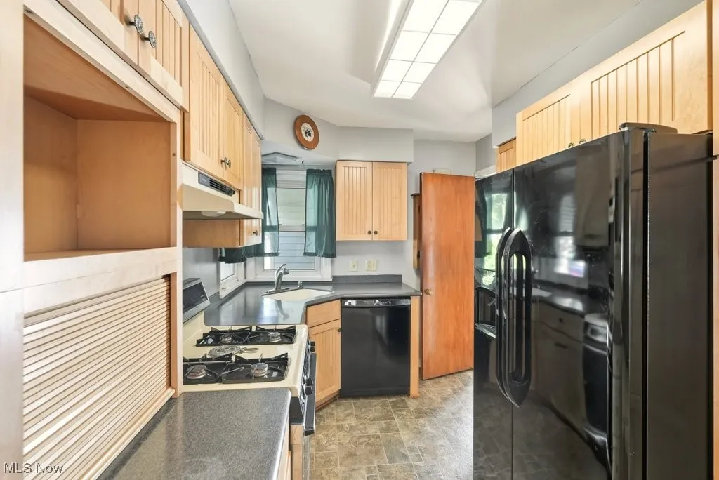 Kitchen featuring black appliances, under cabinet range hood, dark countertops, and light brown cabinetry