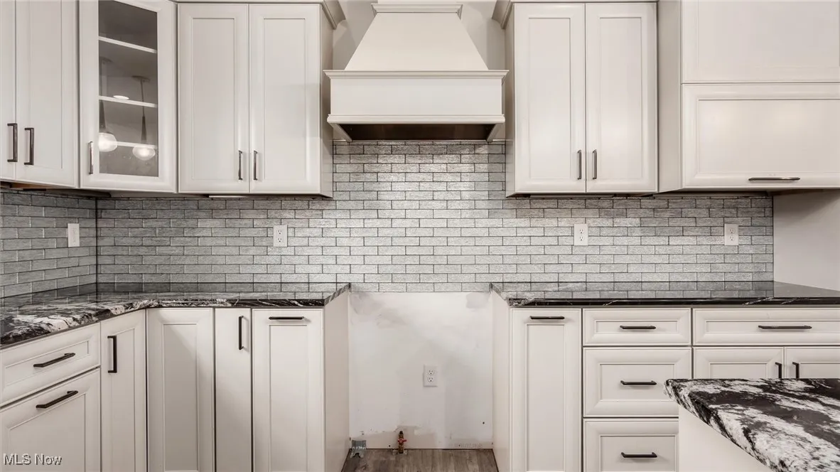 Kitchen featuring custom exhaust hood, backsplash, dark stone counters, and white cabinets