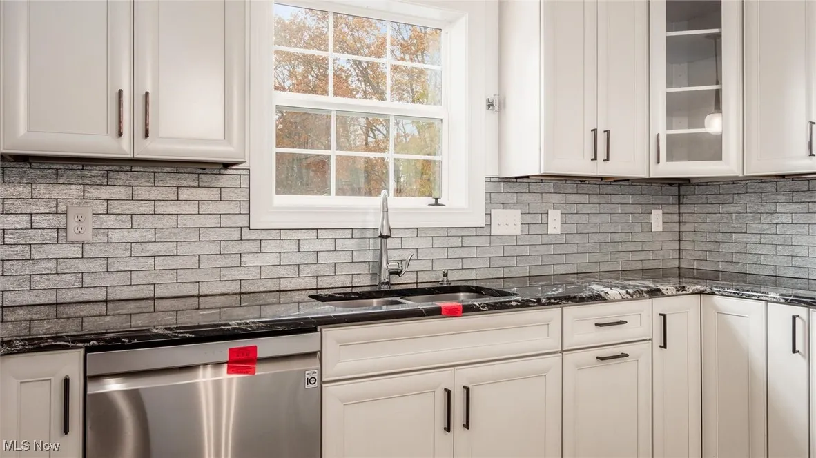 Kitchen featuring dark stone countertops, dishwasher, glass insert cabinets, backsplash, and white cabinets
