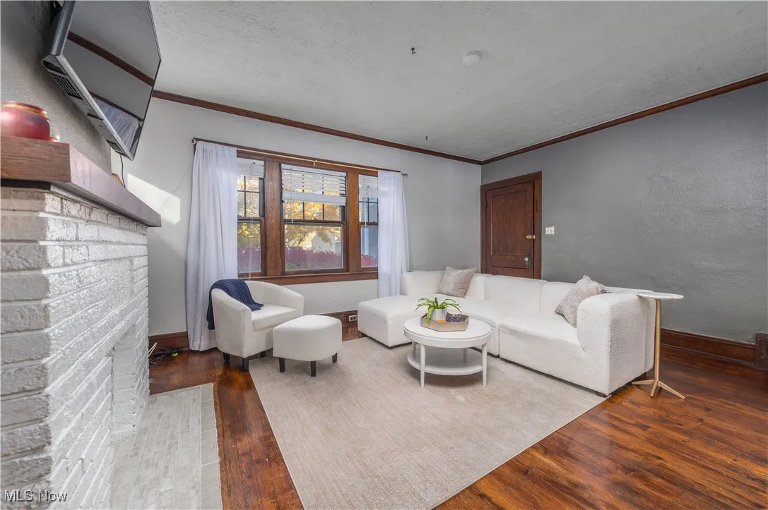 Living room with wood finished floors, a textured wall, a textured ceiling, a brick fireplace, and crown molding