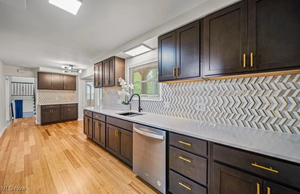 Kitchen with dark brown cabinets, stainless steel dishwasher, light wood-style floors, light stone countertops, and backsplash
