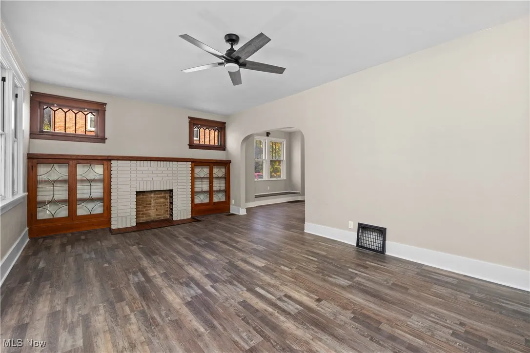 Unfurnished living room with arched walkways, dark wood-style flooring, a brick fireplace, a baseboard radiator, and a ceiling fan