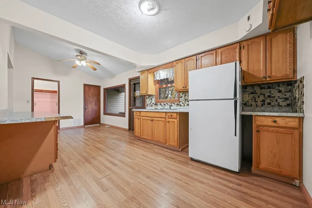 Kitchen featuring freestanding refrigerator, tasteful backsplash, light wood-type flooring, brown cabinets, and a ceiling fan