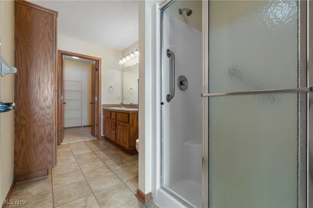 Bright, functional primary bath featuring a true step-in shower with grab bar, double vanity with large mirror, easy-care tile flooring, and a tall linen cabinet—plus direct access to a walk-in closet.