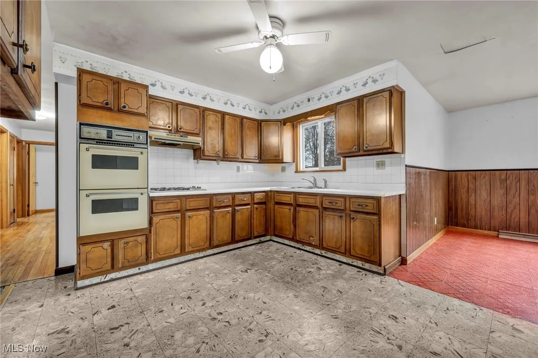 Kitchen featuring brown cabinetry, light countertops, double oven, ceiling fan, and backsplash