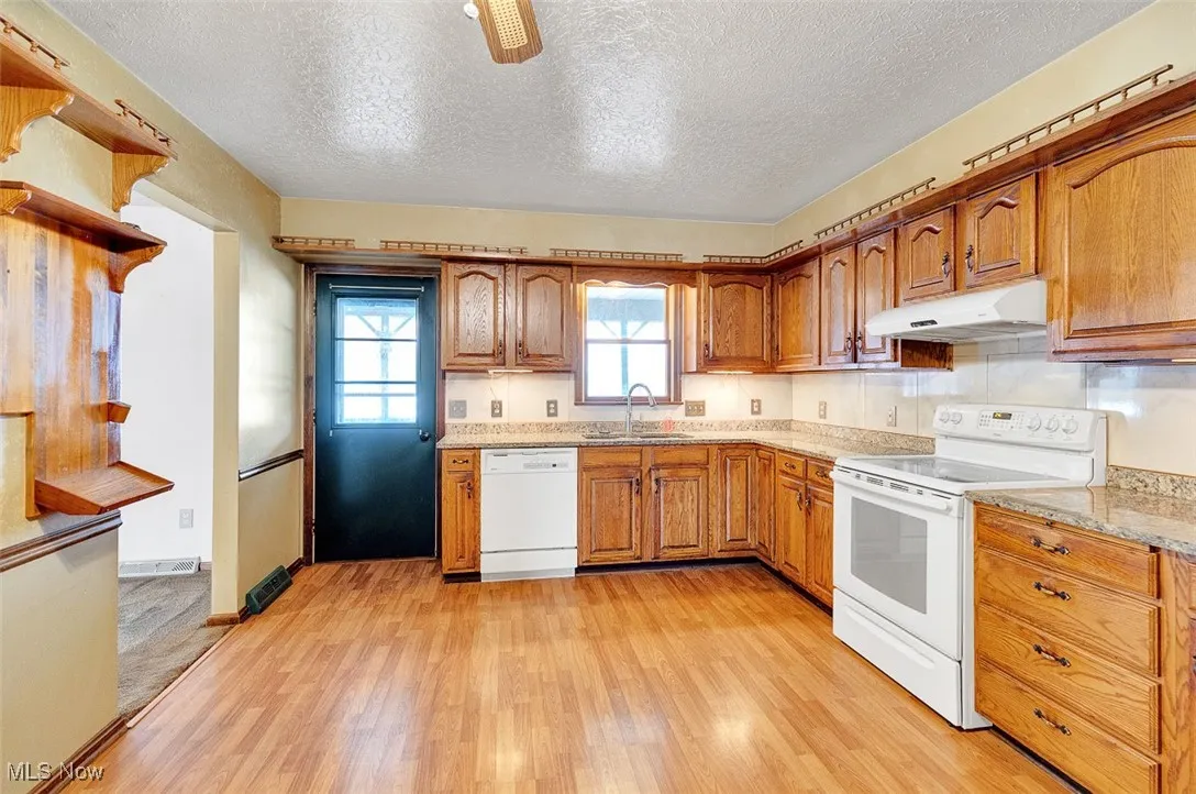 Kitchen with white appliances, a textured ceiling, brown cabinets, light wood-style flooring, and light stone countertops
