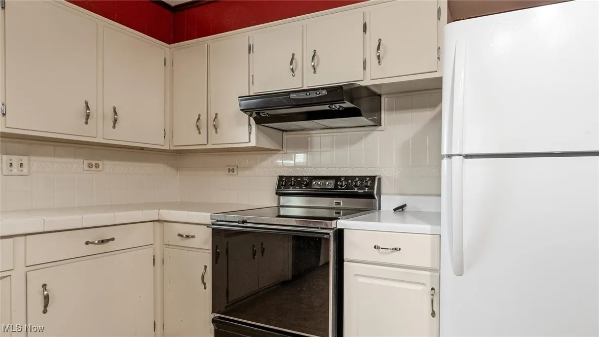Kitchen featuring freestanding refrigerator, black electric range, under cabinet range hood, decorative backsplash, and tile countertops