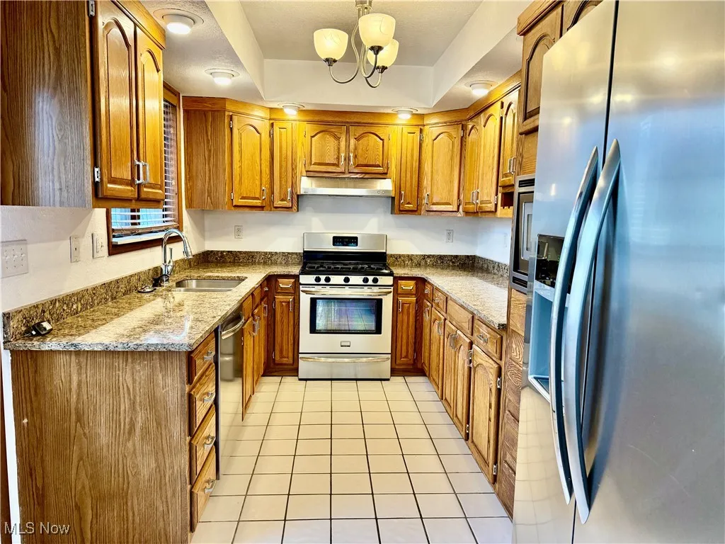 Kitchen with stainless steel appliances, brown cabinetry, light stone counters, light tile patterned floors, and a chandelier