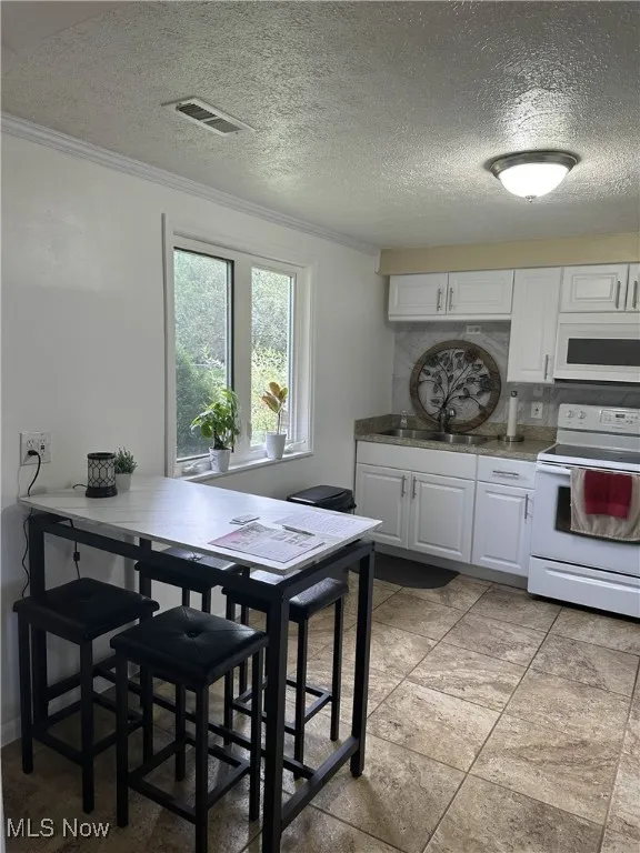 Kitchen featuring white appliances, a textured ceiling, white cabinets, ornamental molding, and a breakfast bar area
