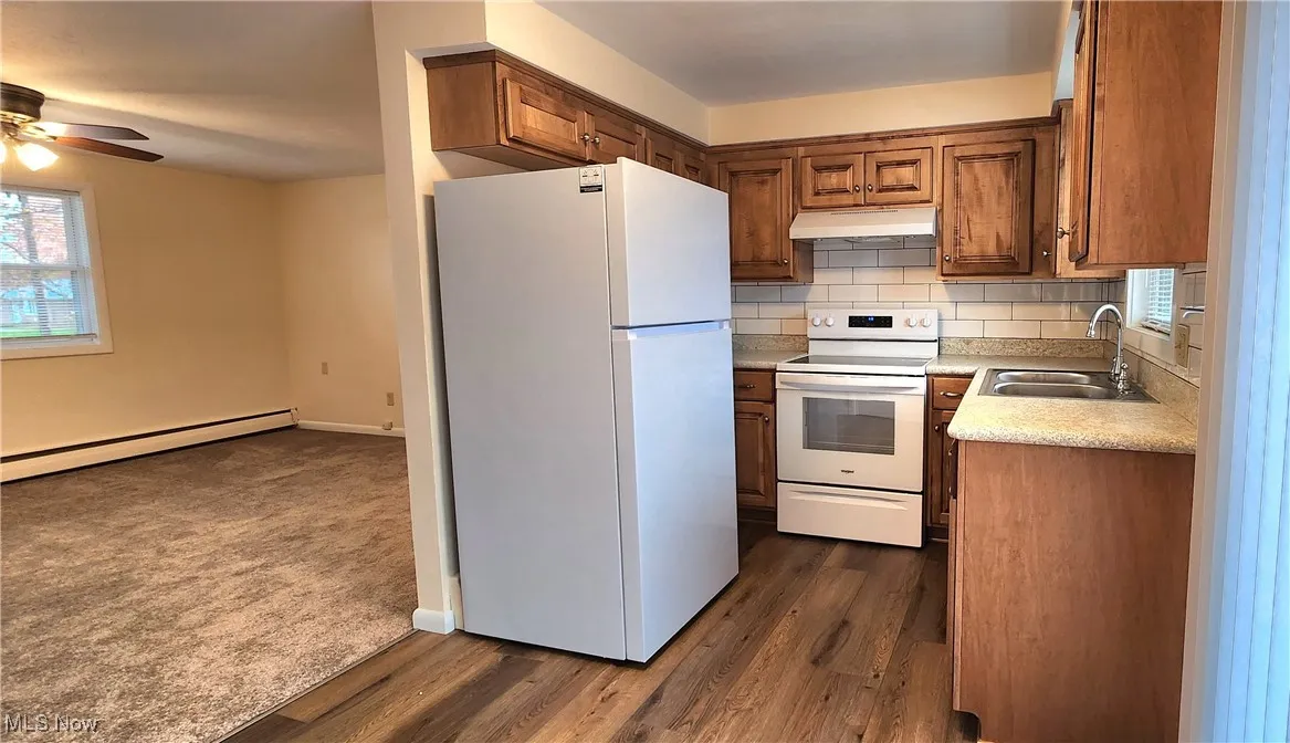 Kitchen featuring white appliances, tasteful backsplash, brown cabinetry, under cabinet range hood, and a baseboard radiator