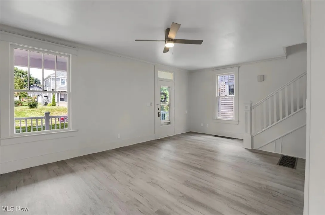 Unfurnished living room with light wood-style flooring, crown molding, a ceiling fan, and stairway