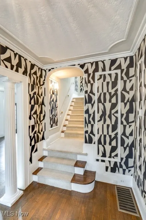 Staircase with wood-type flooring, a textured ceiling, crown molding, arched walkways, and wallpapered walls
