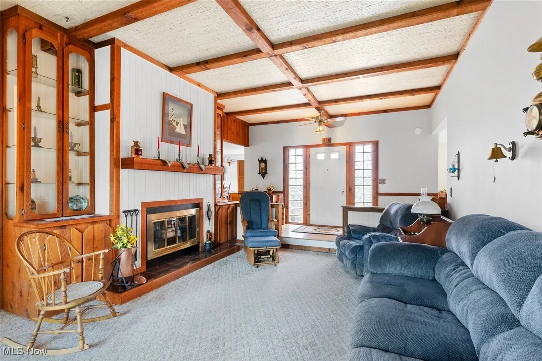 Living area featuring beamed ceiling, a glass covered fireplace, carpet flooring, wooden walls, and a ceiling fan
