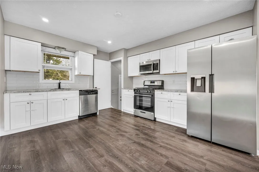 Kitchen with stainless steel appliances, white cabinets, dark wood-style floors, decorative backsplash, and light stone counters