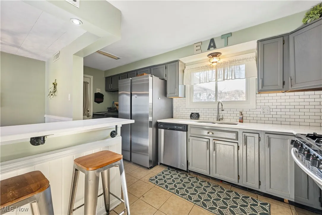Kitchen with gray cabinetry, backsplash, appliances with stainless steel finishes, light tile patterned floors, and a breakfast bar