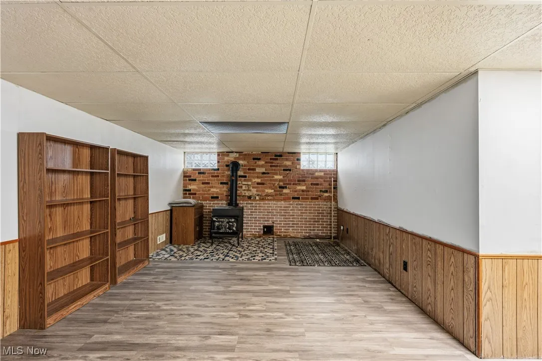 Basement with wainscoting, a drop ceiling, wood walls, a wood stove, and wood finished floors
