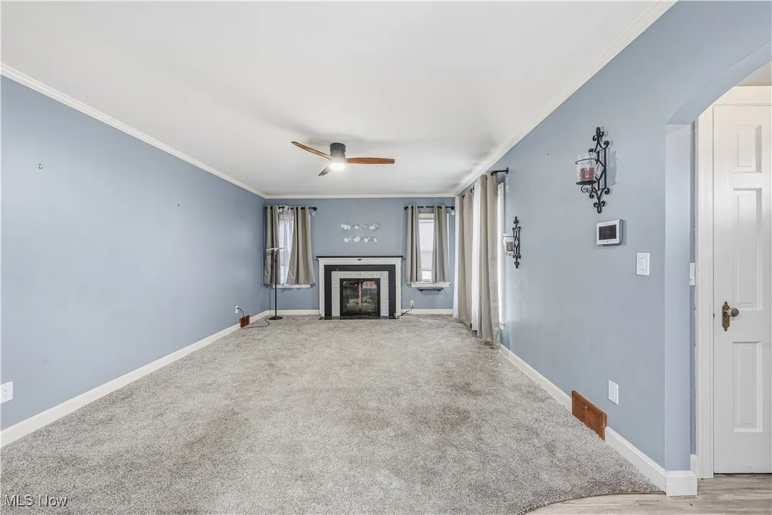Unfurnished living room featuring crown molding, arched walkways, carpet, a ceiling fan, and a fireplace with flush hearth