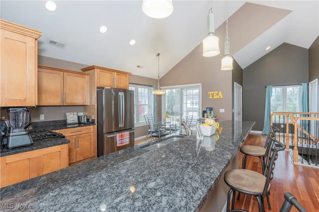 Kitchen with freestanding refrigerator, wood finished floors, hanging light fixtures, high vaulted ceiling, and dark stone counters
