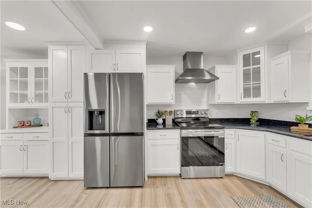 Kitchen with glass insert cabinets, stainless steel appliances, wall chimney range hood, white cabinets, and light wood-type flooring