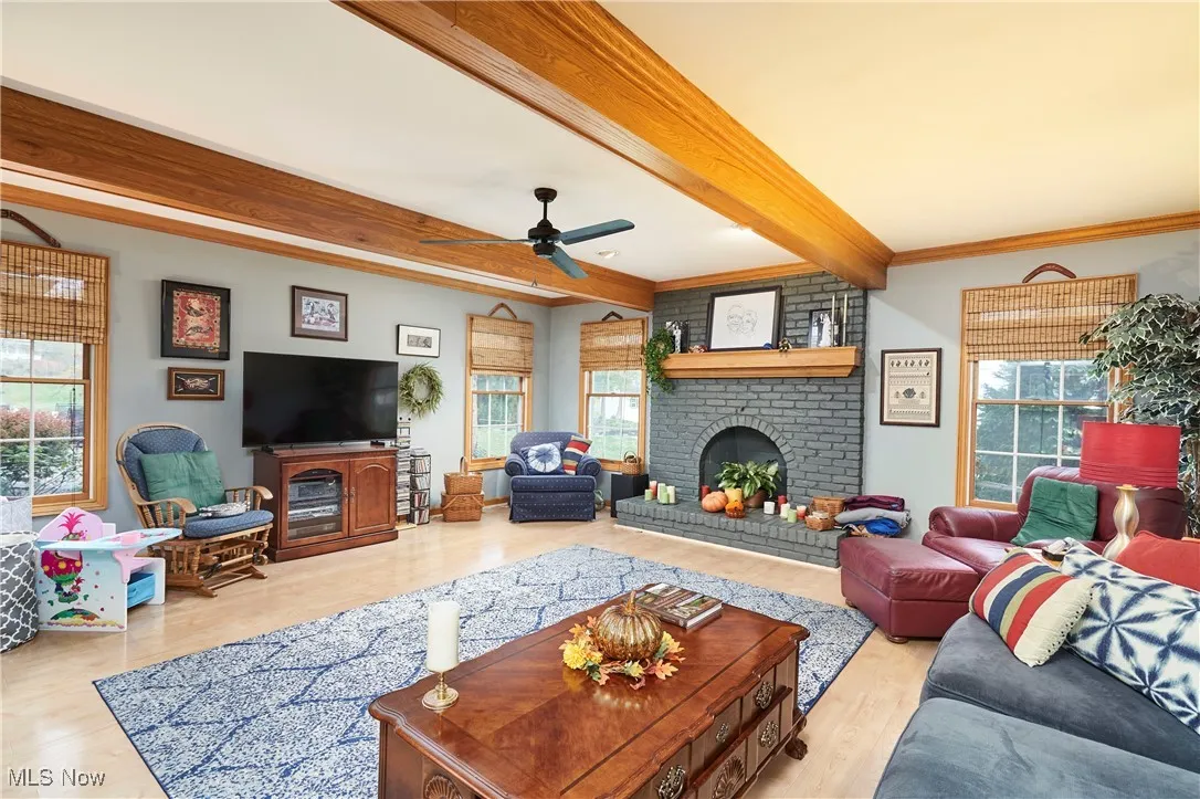 Living room with a fireplace, beam ceiling, light wood-style floors, ceiling fan, and crown molding