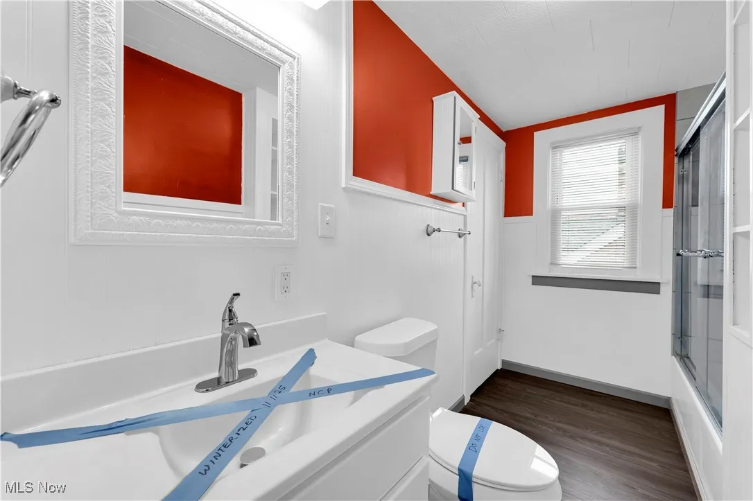 Full bath with vanity, dark wood-type flooring, combined bath / shower with glass door, and wainscoting