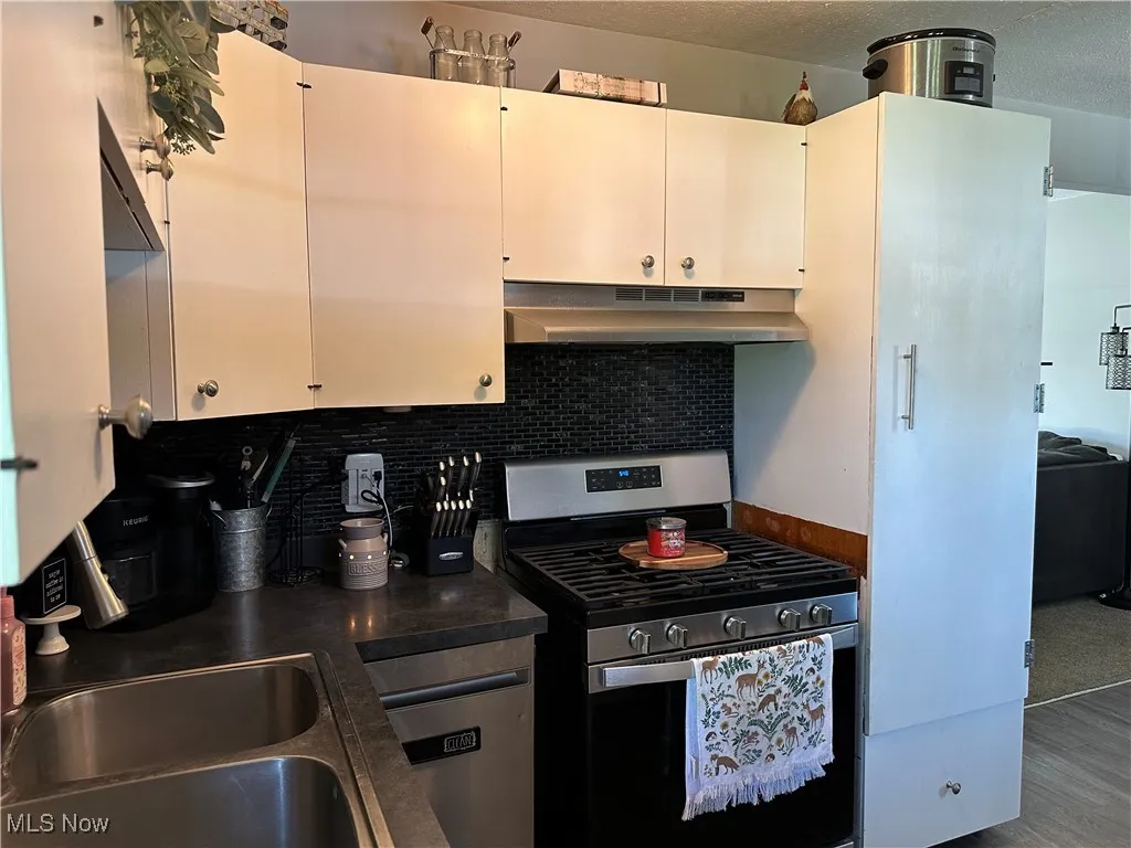Kitchen with dark countertops, stainless steel gas range, under cabinet range hood, wood finished floors, and backsplash