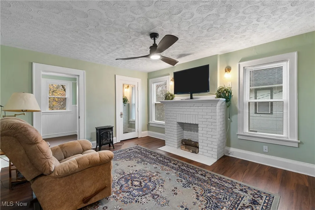Living area featuring dark wood finished floors, a textured ceiling, a brick fireplace, a wood stove, and ceiling fan