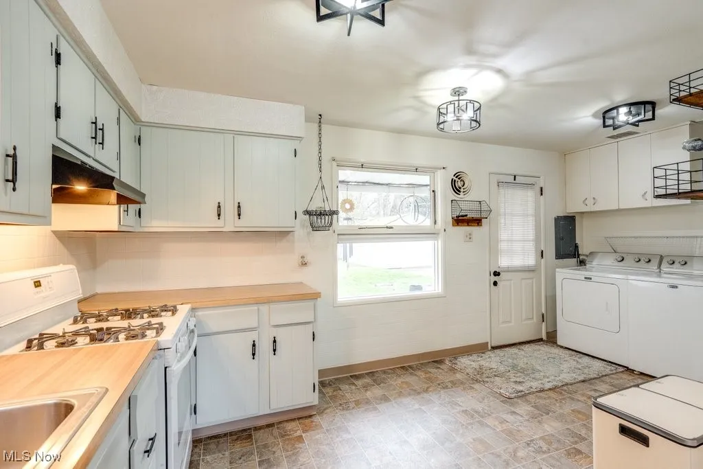 Kitchen with white gas stove, light countertops, under cabinet range hood, washing machine and dryer, and electric panel