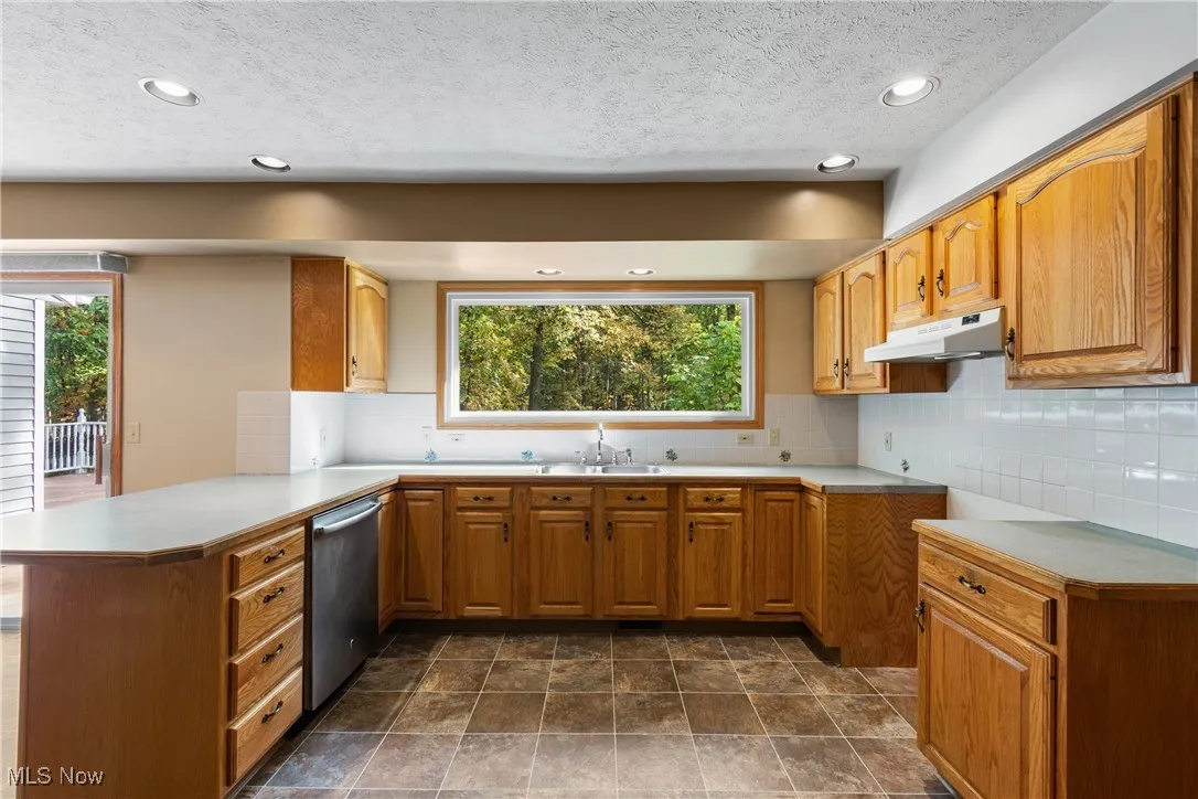 Kitchen with light countertops, decorative backsplash, under cabinet range hood, stainless steel dishwasher, and a textured ceiling
