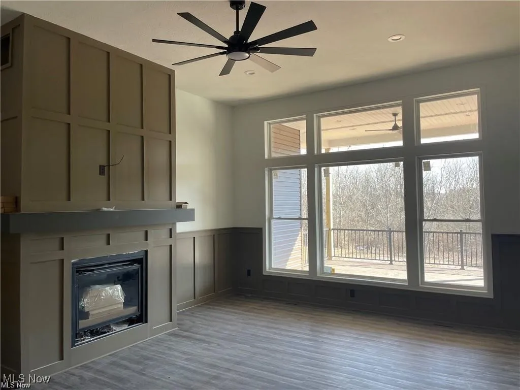 Unfurnished living room with plenty of natural light, a glass covered fireplace, wood finished floors, a ceiling fan, and wainscoting