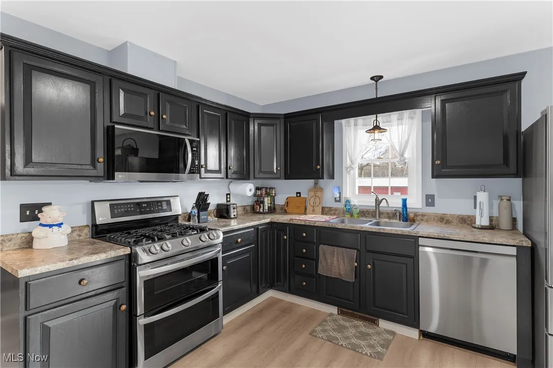 Kitchen with stainless steel appliances, light wood-style flooring, decorative light fixtures, dark cabinetry, and light​​‌​​​​‌​​‌‌​‌‌​​​‌‌​‌​‌​‌​​​‌​​ countertops