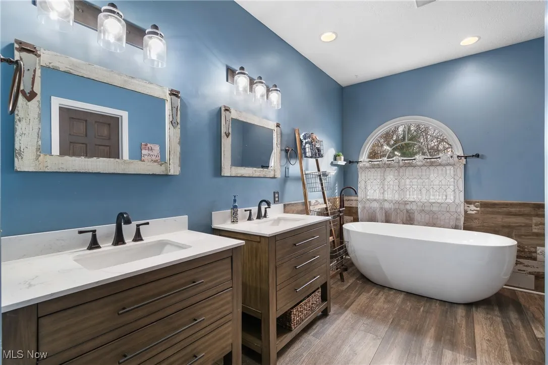 Bathroom featuring two vanities, tile walls, a freestanding bath, a wainscoted wall, and dark wood-style floors