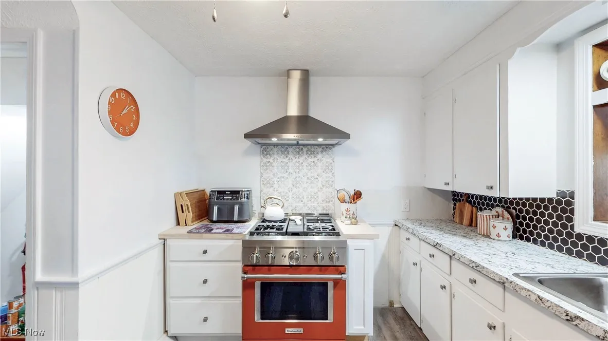 Kitchen featuring decorative backsplash, gas stove, light countertops, white cabinetry, and wall chimney exhaust hood