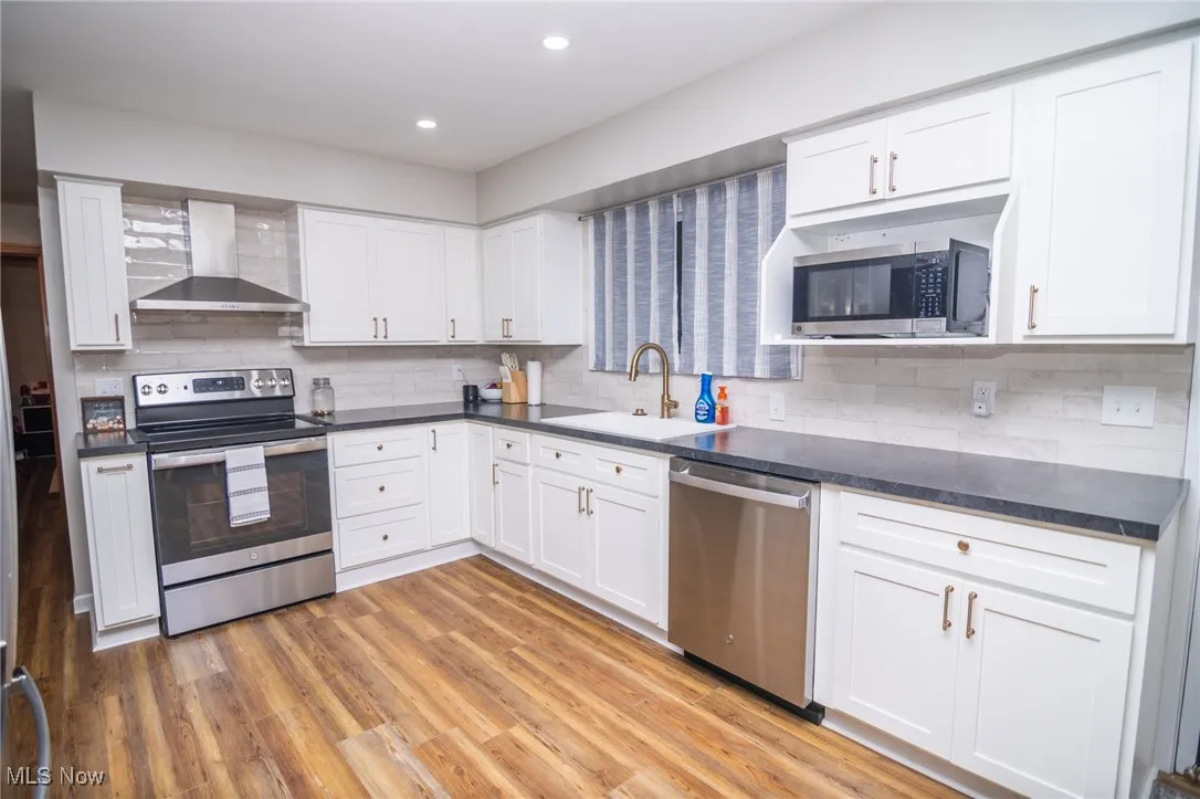 Kitchen with stainless steel appliances, wall chimney exhaust hood, white cabinets, light wood finished floors, and backsplash