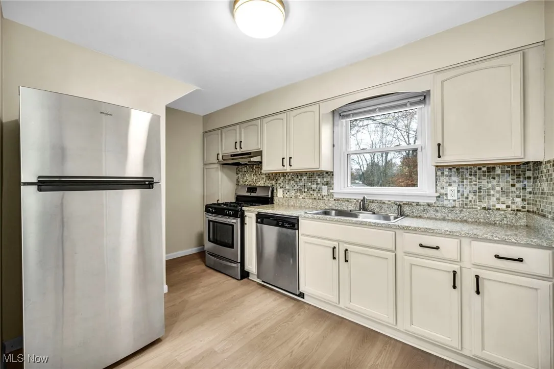 Kitchen featuring appliances with stainless steel finishes, light wood-type flooring, decorative backsplash, under cabinet range hood, and light stone counters
