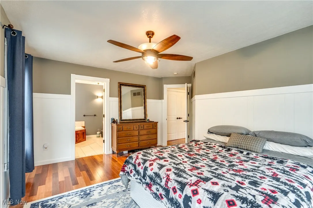 Bedroom with light wood-style flooring, ceiling fan, a wainscoted wall, and connected bathroom
