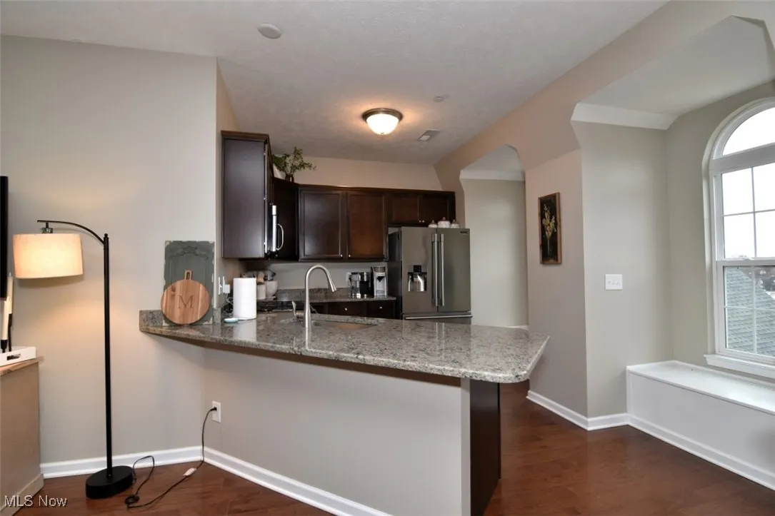 Kitchen with dark brown cabinets, stainless steel fridge, light stone counters, a peninsula, and dark wood finished floors