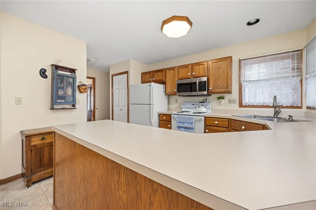 Kitchen with brown cabinetry, light countertops, white appliances, a peninsula, and light tile patterned floors