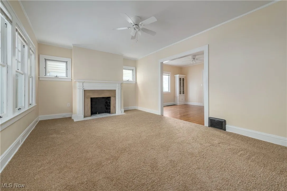 Unfurnished living room featuring ornamental molding, a fireplace with flush hearth, light colored carpet, and ceiling fan
