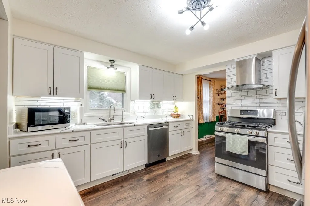 Kitchen with appliances with stainless steel finishes, white cabinetry, wall chimney range hood, dark wood-style flooring, and a textured ceiling