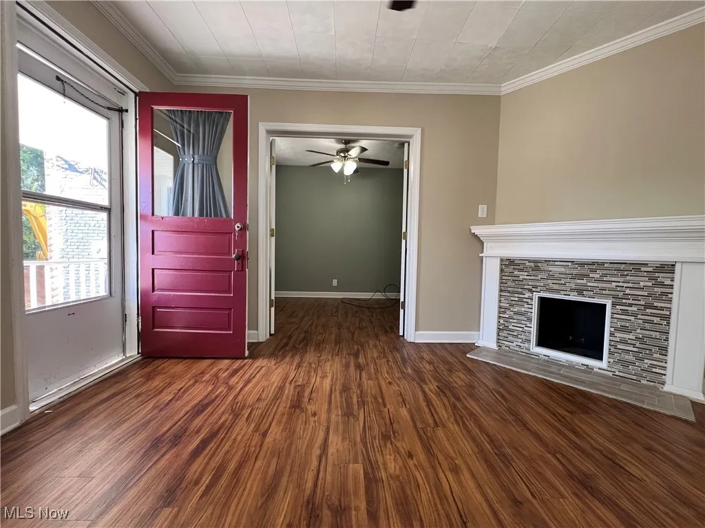 Unfurnished living room featuring dark wood finished floors, a stone fireplace, ceiling fan, and crown molding